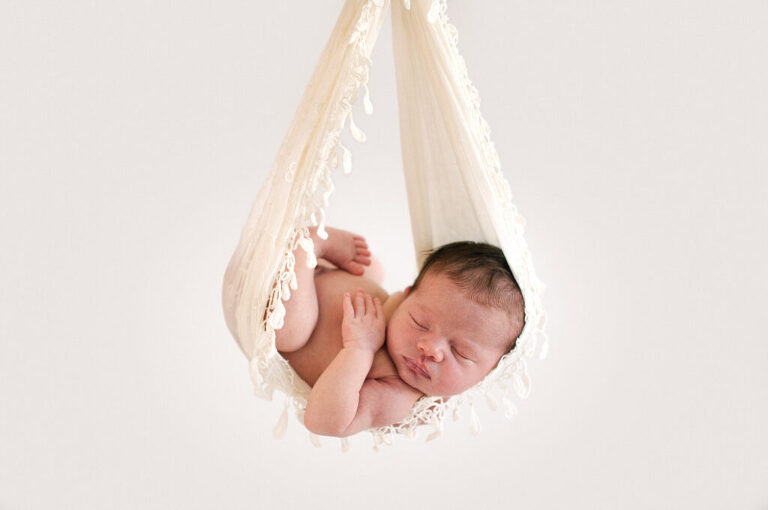 A newborn baby sleeping and laying on her back cradled in a beautiful white fabric with a white backdrop