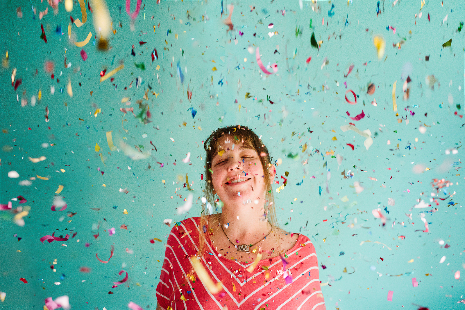 Self portrait of photographer Lindsay smiling joyfully as confetti falls around her in front of a turquoise backdrop
