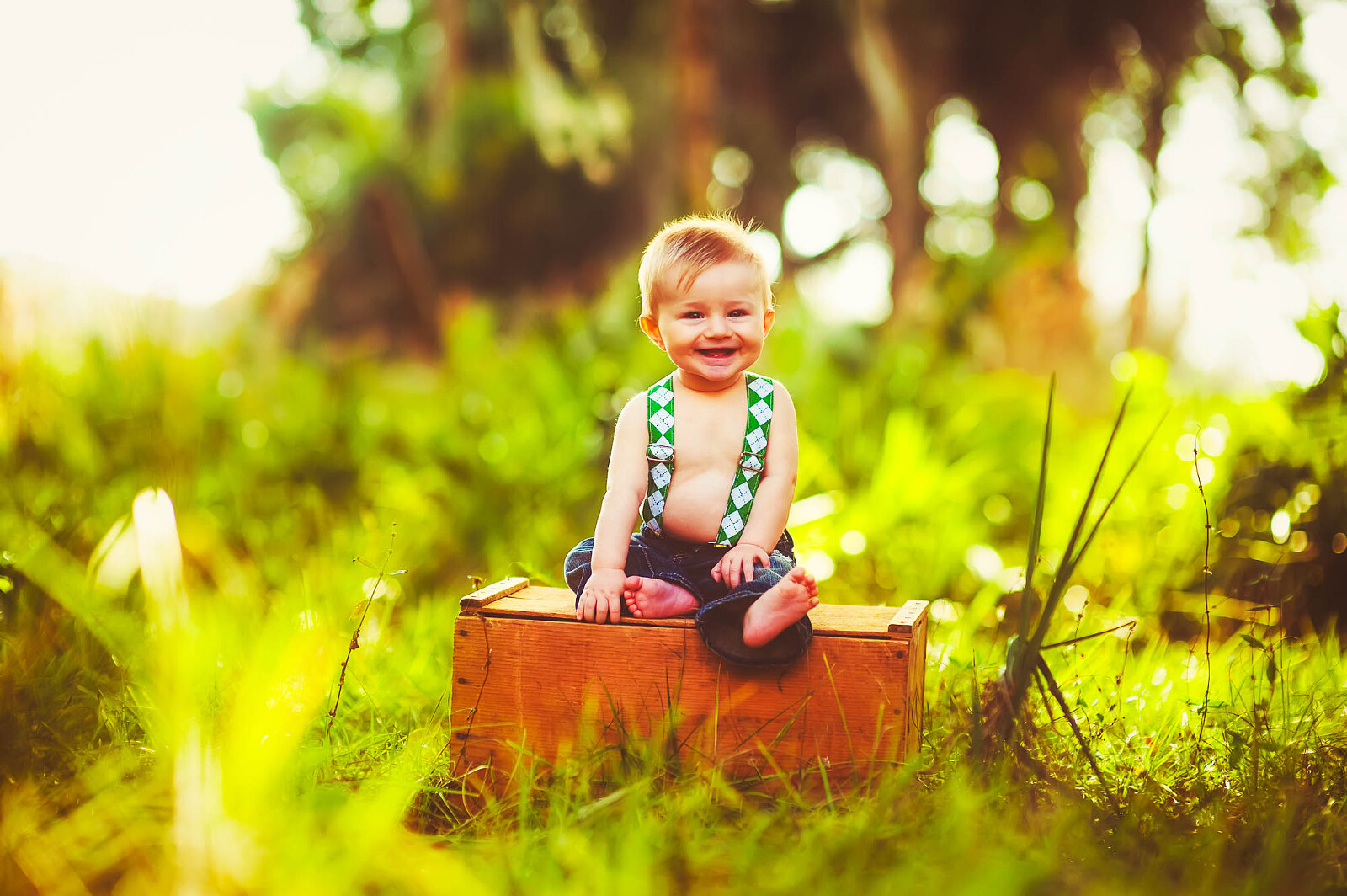 Smiling baby boy sitting on a wooden crate in sunlit field with greenery
