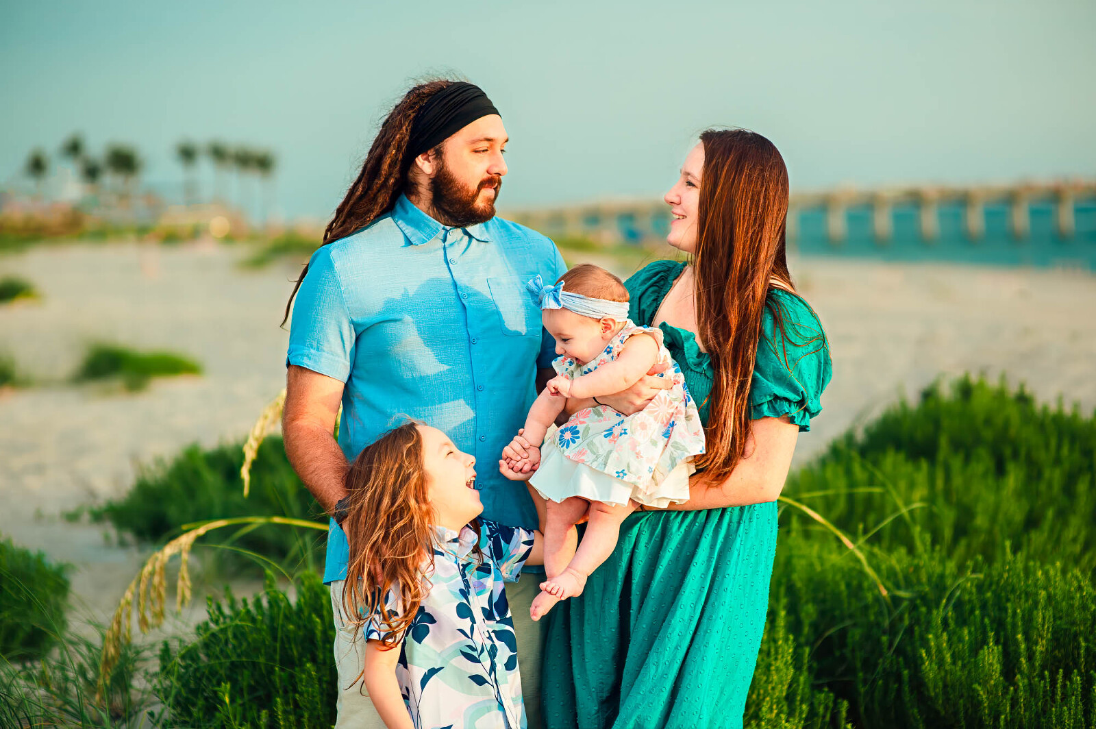 Sarasota family of four with a baby girl and young son, standing near the beach with greenery and pier in background
