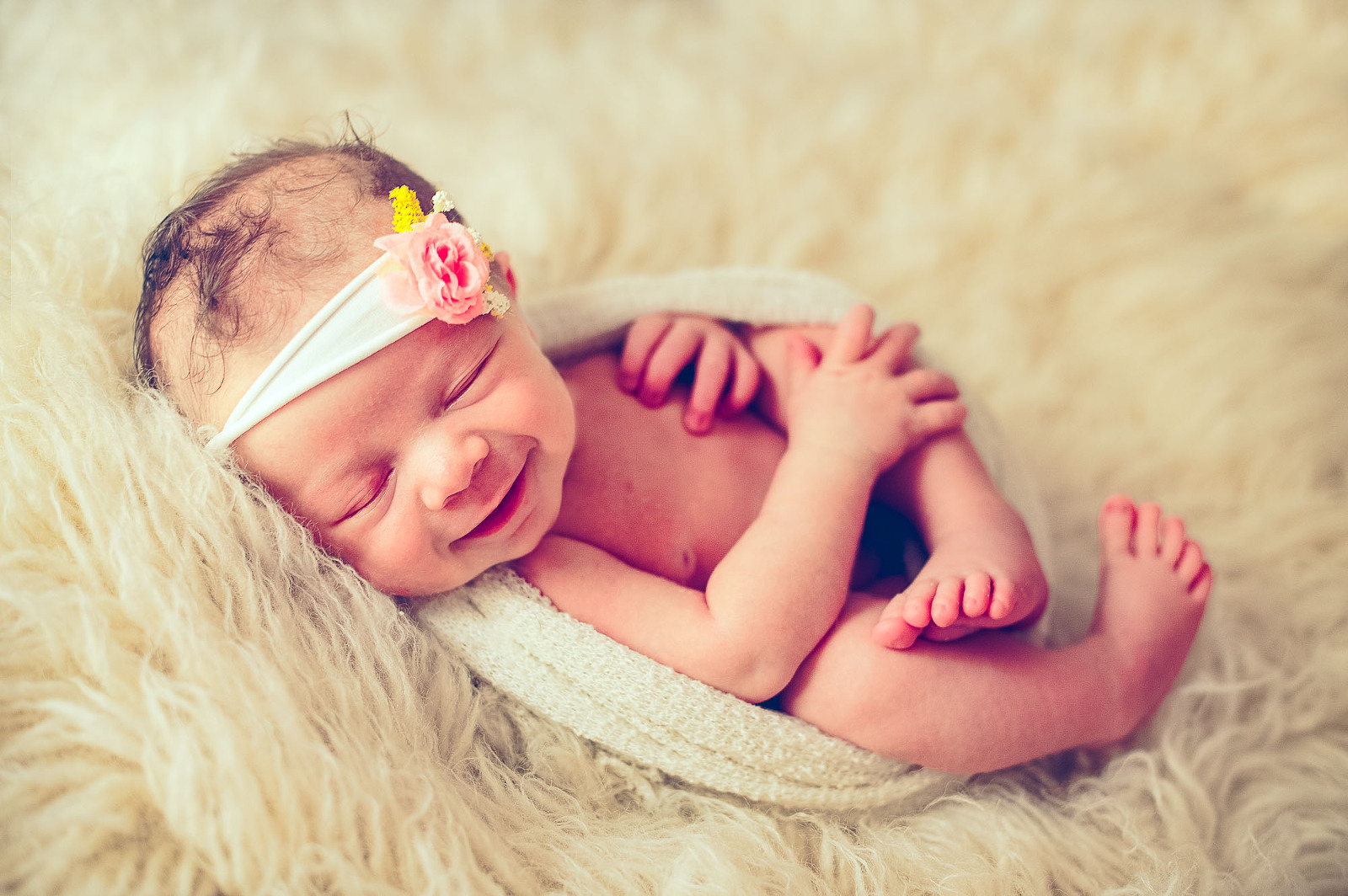 Newborn baby girl smiling in a cozy knit wrap with a floral headband on a soft fur blanket