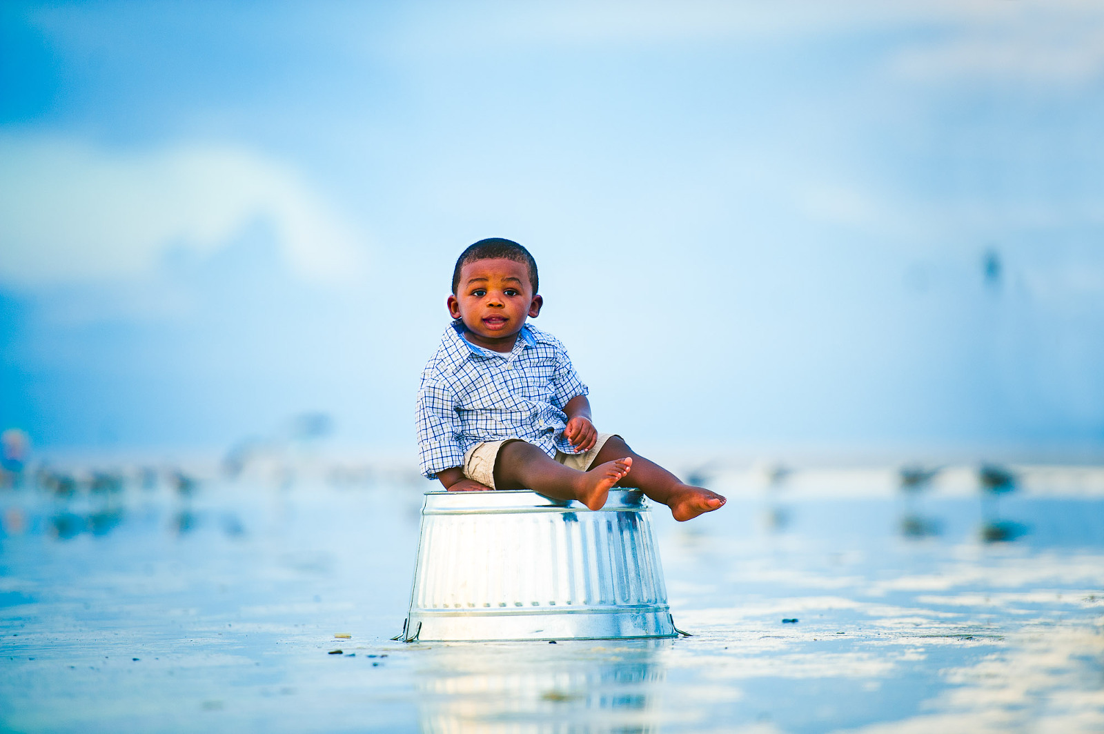 Young boy sitting on a metal pail in the shallow water during sunset at the beach