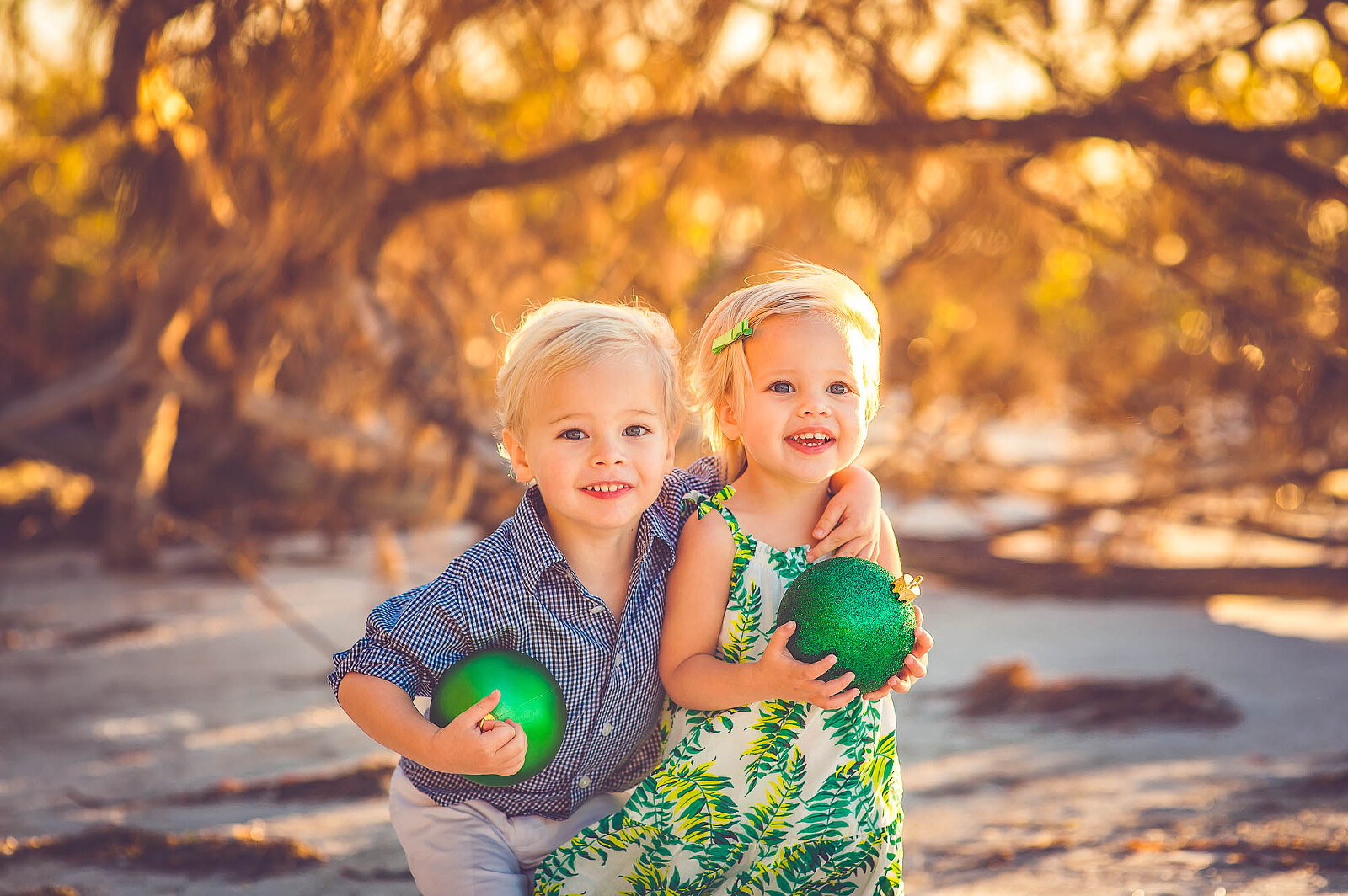 Blonde toddler siblings smiling and holding green ornaments during a holiday-themed beach photo session in Sarasota, Florida