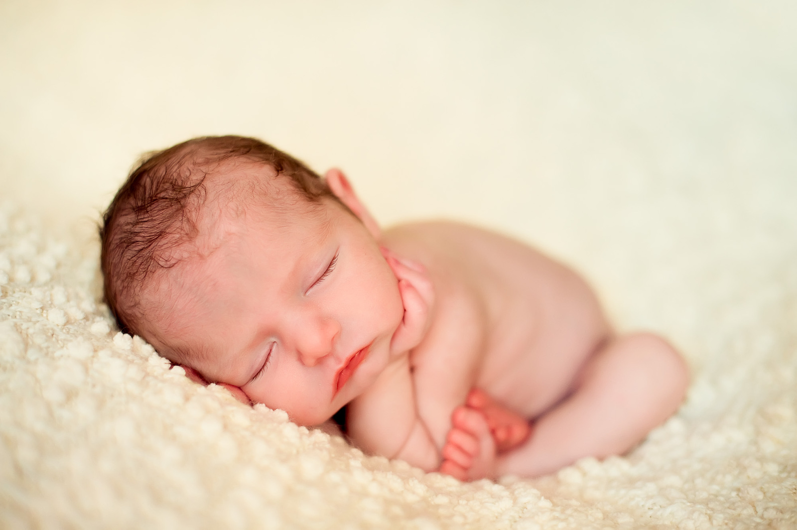 Newborn baby peacefully sleeping on a soft, ivory textured blanket with hands tucked under cheek