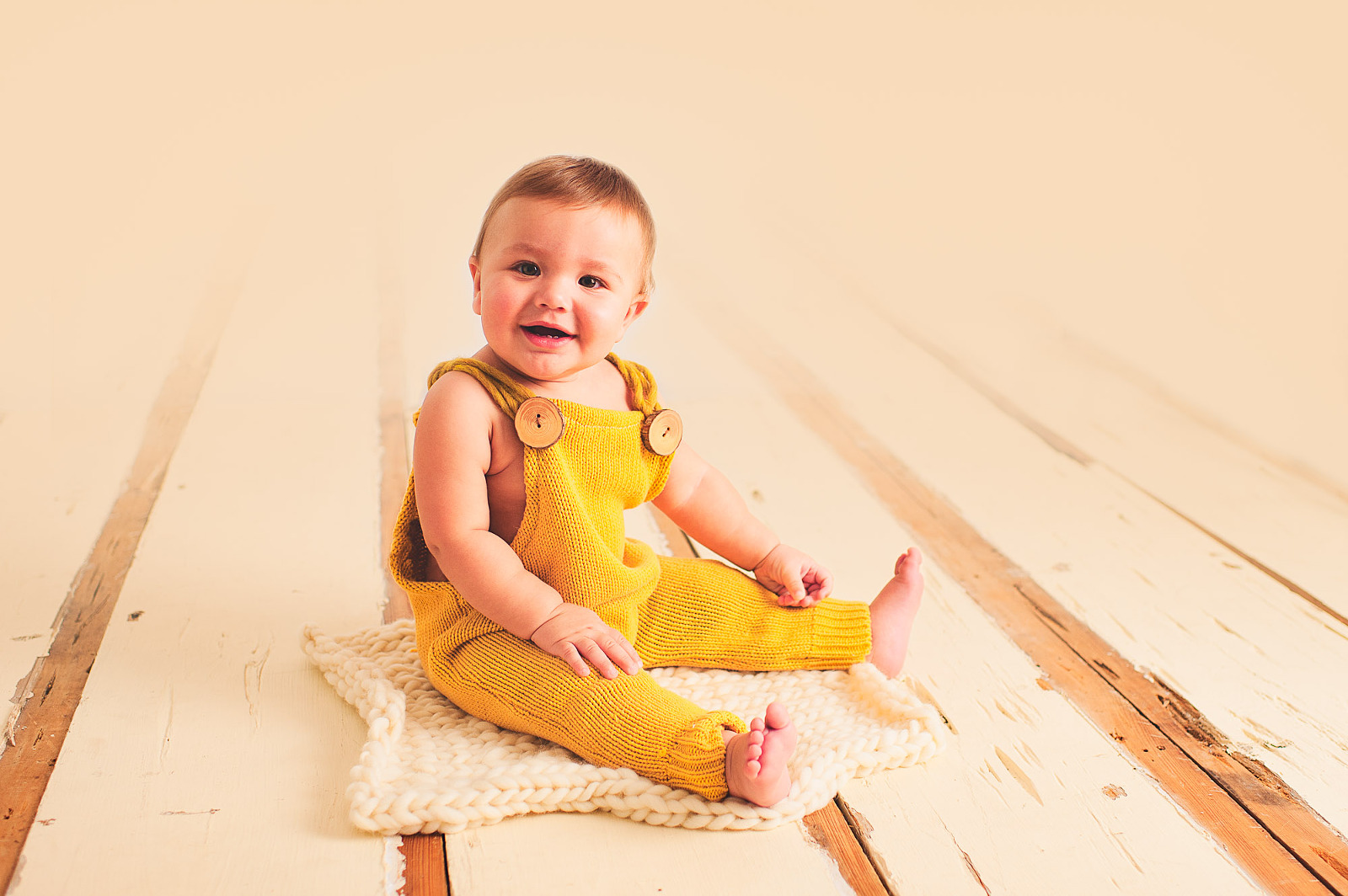 Smiling baby boy in a mustard yellow romper sitting on a white wood floor in a Sarasota studio